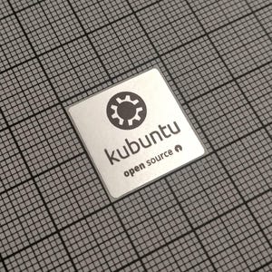 May include: A square, silver sticker with the word "kubuntu" in a sans-serif font, and the words "open source" below it. The sticker has a black gear icon above the word "kubuntu". The sticker is on a gray grid background.