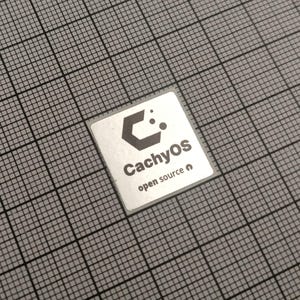 May include: A small, square silver sticker with the text "CachyOS open source" and a stylized logo. The sticker is set against a grey grid background.