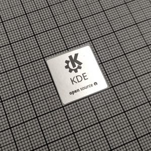May include: A square, silver sticker featuring the KDE logo, a gear-like design with a stylized 'K' in the center, and the text 'KDE open source'. The sticker is on a grey grid background.