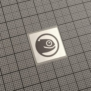 May include: A square sticker featuring a grey fish icon within a circle. The sticker has a white border and is placed on a grey grid background. The fish icon is a simple, stylised design.