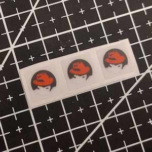 May include: Three small square stickers arranged in a horizontal row. Each sticker features a circular graphic of a person wearing a bright orange fedora hat against a dark gray background. The stickers are printed on a clear adhesive backing.