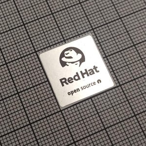 Può includere: Un adesivo quadrato argentato con il logo Red Hat, raffigurante la silhouette di una persona con un fedora. Il testo "Red Hat" è sotto il logo, con "open source" sotto. L'adesivo è su uno sfondo a griglia grigia.