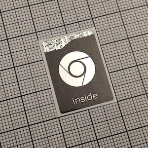 Könnte beinhalten: Ein rechteckiger schwarzer Aufkleber mit dem weißen Chrome-Logo, einem kreisförmigen Design mit einem zentralen Punkt und drei gebogenen Linien. Das Wort "inside" ist weiß unter dem Logo aufgedruckt. Der Aufkleber befindet sich auf einem grauen Gitterhintergrund.