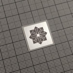 May include: A square, silver sticker with a geometric starburst design in dark gray. The sticker is centered on a gray grid background, creating a contrasting visual effect. The design is symmetrical and modern.