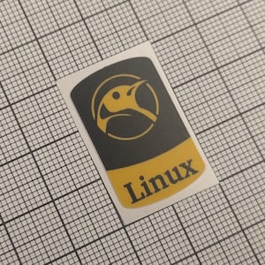 Puede incluir: Adhesivo rectangular con el logotipo de Linux. El adhesivo tiene un fondo negro con un logotipo de pájaro amarillo dentro de un círculo. La palabra "Linux" está impresa en amarillo en la parte inferior.