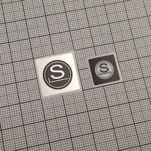 Peut inclure: Deux autocollants carrés avec un motif noir et blanc. Chaque autocollant présente la lettre "S" blanche à l'intérieur d'un cercle noir, avec un trait horizontal en dessous. L'un est argenté, l'autre est noir.