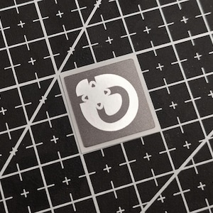 May include: A square sticker with a silver design on a gray background. The design is a stylized, abstract shape with a circular element and a few cut-out sections. The sticker is placed on a black cutting mat with white grid lines.