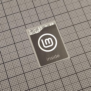 Könnte beinhalten: Ein rechteckiger, dunkelgrauer Aufkleber mit dem Text "inside" unterhalb eines weißen kreisförmigen Logos mit den Buchstaben "lm" darin. Der Aufkleber hat eine teilweise zerrissene Oberkante und befindet sich vor einem grauen Rasterhintergrund.