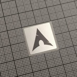 May include: A square sticker with a silver border and a gray logo. The logo is an abstract design featuring a stylized arrow shape with a curved opening at the bottom. The sticker is on a gray grid background.