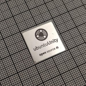 Puede incluir: Un pequeño componente cuadrado de color plateado con el texto "ubuntuUnity" y "open source" impreso. El componente tiene un logotipo circular con tres formas interconectadas. El fondo es una cuadrícula gris.