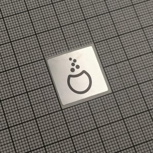 Op de afbeelding: Een kleine, vierkante, zilveren sticker met een zwarte afbeelding van een beker met bubbels die eruit komen. De sticker staat op een grijze rasterachtergrond.