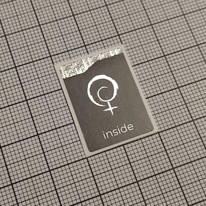 May include: A rectangular sticker with a grey background and a white border. The sticker features a white symbol and the word "inside" in white text. The sticker is placed on a grey grid.
