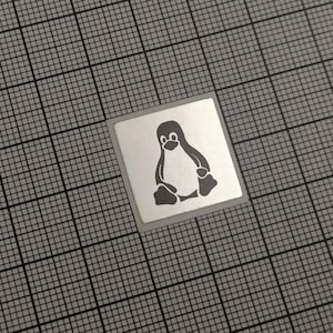 Könnte beinhalten: Ein quadratischer, silberner Aufkleber mit dem Linux-Maskottchen Tux, dem Pinguin, in Dunkelgrau. Der Aufkleber befindet sich vor einem grauen Rasterhintergrund, was einen visuellen Kontrast erzeugt.