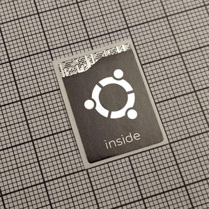 Pode incluir: Um autocolante a preto e branco com o logótipo Ubuntu, um círculo com três pontos e a palavra "inside". O autocolante está sobre um fundo de grelha cinzenta.