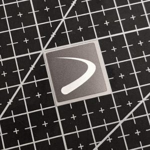 May include: A square sticker with a gray background and a white curved design. The sticker is on a black cutting mat with white grid lines and plus signs.