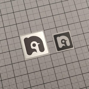 May include: Two square stickers with a black and white graphic of a stylized bird. The larger sticker has a silver border. The stickers are on a grid background.