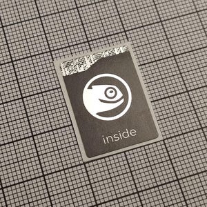 Op de afbeelding: Een rechthoekige, donkergrijze sticker met een wit logo van een gestileerde vis in een cirkel. Het woord "inside" staat eronder in het wit gedrukt. De sticker ligt op een grijze rasterachtergrond.