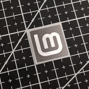 May include: A square sticker with a gray background and a white logo featuring the letters "l" and "m" in a stylized design. The sticker is on a black cutting mat with white grid lines.