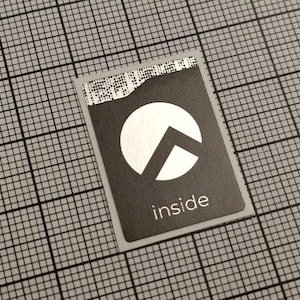 Könnte beinhalten: Ein rechteckiger schwarz-weißer Aufkleber mit dem weißen Aufdruck "inside". Der Aufkleber zeigt einen weißen Kreis mit einem schwarzen Pfeil nach oben. Der Aufkleber befindet sich auf einem Rasterhintergrund.