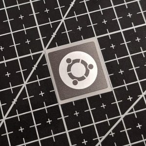 Może przedstawiać: Kwadratowa naklejka z logo Ubuntu, stylizowanym białym okręgiem z trzema kropkami i centralnym pierścieniem. Naklejka ma szarą obwódkę i jest umieszczona na czarnej macie do cięcia z białymi liniami siatki i znakami plus.
