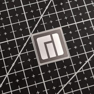 May include: A square, silver sticker with a gray border and a white, abstract design. The sticker is on a black cutting mat with white grid lines and diagonal markings.