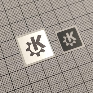 Könnte beinhalten: Zwei quadratische Aufkleber mit einem Zahnrad und dem Buchstaben "K". Einer ist silberfarben mit schwarzem Design, der andere schwarz mit weißem Design. Sie befinden sich auf einem grauen Rasterhintergrund.