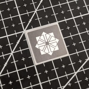 May include: A square sticker with a geometric starburst design in white on a gray background. The sticker is centered on a black cutting mat with white grid lines.