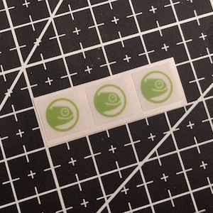 May include: Three square stickers arranged in a horizontal row, each featuring a light green circular icon with a stylised chameleon head profile inside. The stickers are white with a thin border and are positioned on a black grid cutting mat.