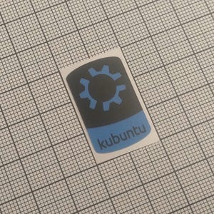 May include: A sticker featuring the Kubuntu logo. The design includes a blue gear icon on a black background, with a blue bar at the bottom containing the word "kubuntu". The sticker is on a grid background.