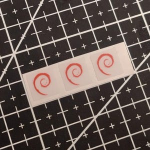 May include: A strip of three small, square, translucent stickers, each featuring a red spiral graphic. The stickers are arranged horizontally in a row, showcasing a simple, clean design with a distinct red swirl pattern on a clear, adhesive backing.