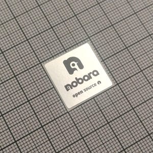 May include: A square, silver sticker with the word "nobara" in a stylized font and the words "open source" below it. The sticker has a gray logo of a bird. The sticker is on a gray grid background.