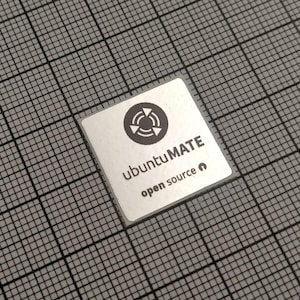 May include: A small, square, silver-colored computer component with the text "ubuntu MATE open source" printed on it. The component has a black graphic of three arrows in a circle. The background is a gray grid.