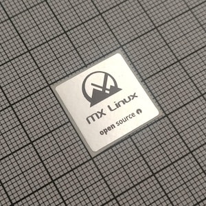 May include: A square, silver sticker with the text "MX Linux open source" in a sans-serif font. The sticker features a stylized mountain and arrow logo. The background is a gray grid.