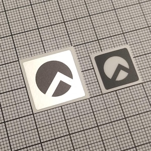May include: Two square stickers with a geometric design. One sticker has a white background with a dark gray circle and a white chevron. The other sticker has a dark gray background with a white circle and chevron.