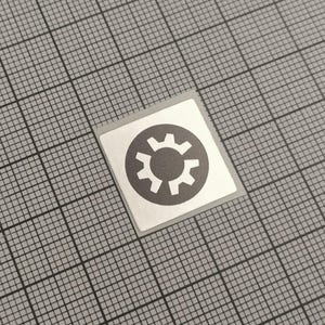 May include: A square sticker with a silver border and a gray gear icon in the center. The gear is inside a gray circle. The sticker is on a gray grid background.