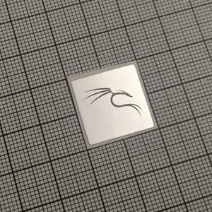 May include: A small, square sticker with a silver border and a black dragon logo. The sticker is centered on a gray grid background.