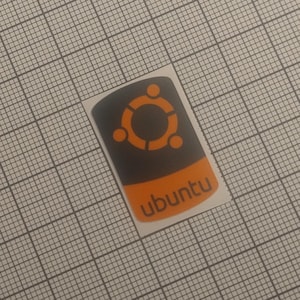 May include: A rectangular sticker with the Ubuntu logo. The top section is black with an orange circular design, and the bottom is orange with the word "ubuntu" in white. The sticker is on a grid background.
