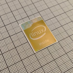 Peut inclure: Un autocollant doré avec le texte "linux inside" et le logo Intel. L'autocollant est rectangulaire avec des coins arrondis et se trouve sur un fond quadrillé.