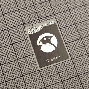 Pode incluir: Um autocolante retangular cinza escuro com um logótipo de pinguim branco dentro de um círculo branco. A palavra "inside" está impressa em branco abaixo do logótipo. O autocolante está sobre um fundo de grelha cinza.