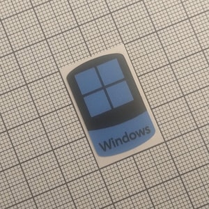 Peut inclure: Un autocollant avec le logo Windows. Le design comprend un rectangle noir avec quatre carrés bleus et une bannière bleue en bas avec le mot "Windows" en blanc.