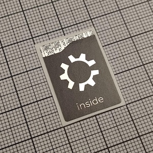 Könnte beinhalten: Ein rechteckiger Aufkleber mit einem weißen Zahnradsymbol und dem Wort "inside" auf dunklem grauem Hintergrund. Der Aufkleber hat eine eingerissene Kante oben und befindet sich vor einem grauen Rasterhintergrund.