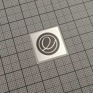 Könnte beinhalten: Ein quadratischer Aufkleber mit silbernem Rand und einem grauen kreisförmigen Logo. Das Logo zeigt ein stilisiertes "e" in einem größeren Kreis. Der Aufkleber befindet sich auf einem grauen Rasterhintergrund.