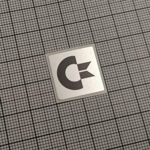Könnte beinhalten: Ein quadratischer Aufkleber mit dem Commodore-Logo, einem stilisierten 'C' mit horizontalem Pfeil. Das Logo ist dunkelgrau auf weißem Grund, mit silbernem Rand. Der Aufkleber befindet sich auf einem grauen Rasterhintergrund.