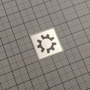 Op de afbeelding: Een vierkante sticker met een grijs tandwielpictogram op een witte achtergrond. Het tandwiel heeft een centrale cirkel en omringende tandwielen. De sticker is geplaatst op een grijze rasterachtergrond.
