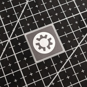 Peut inclure: Un autocollant carré avec un symbole d'engrenage blanc à l'intérieur d'un cercle blanc, sur un fond gris. L'autocollant est posé sur un tapis de coupe noir avec un motif quadrillé et des croix blanches.