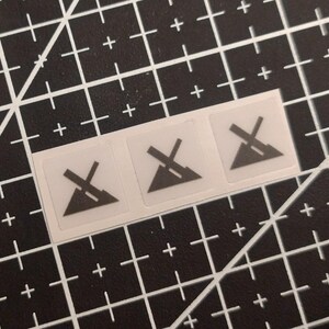 Op de afbeelding: Drie kleine, vierkante, half-transparante stickers in een horizontale rij. Elke sticker bevat een donkergrijs grafisch icoon van een driehoek met een diagonale lijn erdoorheen, vergelijkbaar met het wassymbool voor niet strijken.