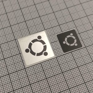 May include: Two square stickers with a black and white Ubuntu logo. The larger sticker is silver, and the smaller one is black. The Ubuntu logo features a circle with three smaller circles around it.