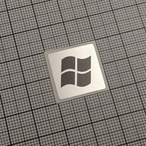 Könnte beinhalten: Ein quadratischer Aufkleber mit silbernem Rand und einem grauen Windows-Logo. Das Logo zeigt vier gebogene Rechtecke, die einem Fenster ähneln. Der Aufkleber befindet sich auf einem grauen Gitterhintergrund.