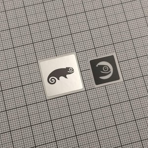 May include: Two square stickers. One is silver with a black chameleon silhouette. The other is black with a white eye design. Both stickers are on a grey grid background.