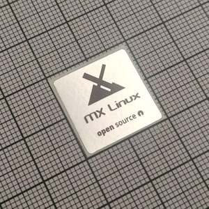 May include: A square, silver sticker with the text "MX Linux open source" in a sans-serif font. The logo features a stylized X and a triangle. The sticker is on a gray grid background.